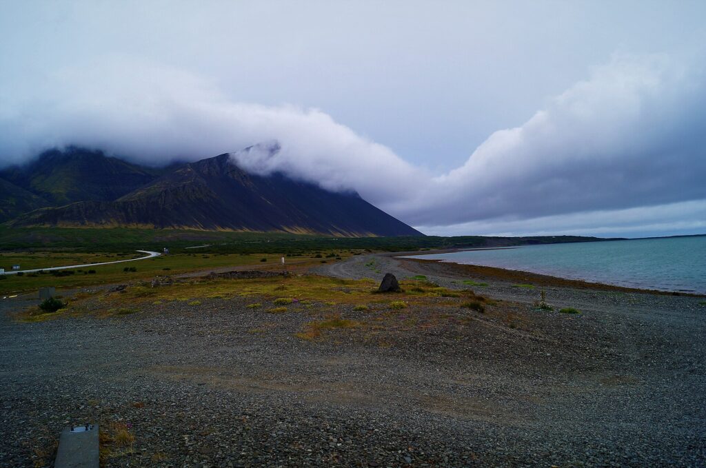 A paved road running along the grassy shore of Borgarfjörður at the intersection of Road 1 and Road 50 in West Iceland, with low hills and water in the background under a cloudy sky.