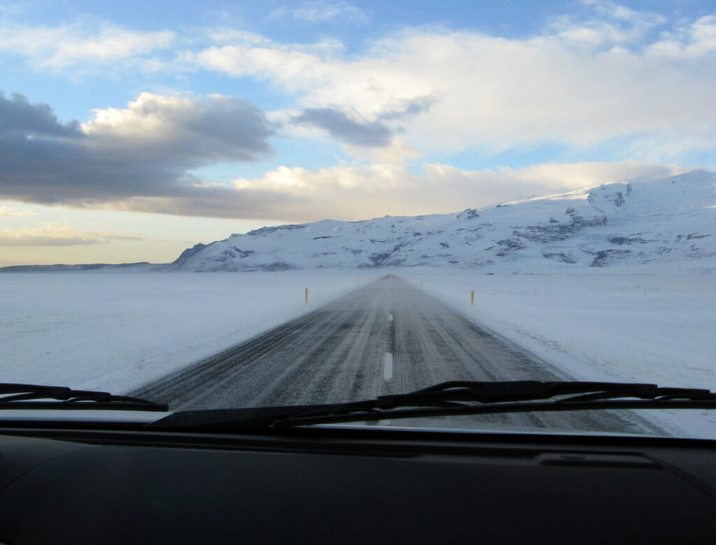 An ice- and snow-covered stretch of Iceland's Ring Road (Route 1) in November, running toward distant snowy mountains under an overcast sky.