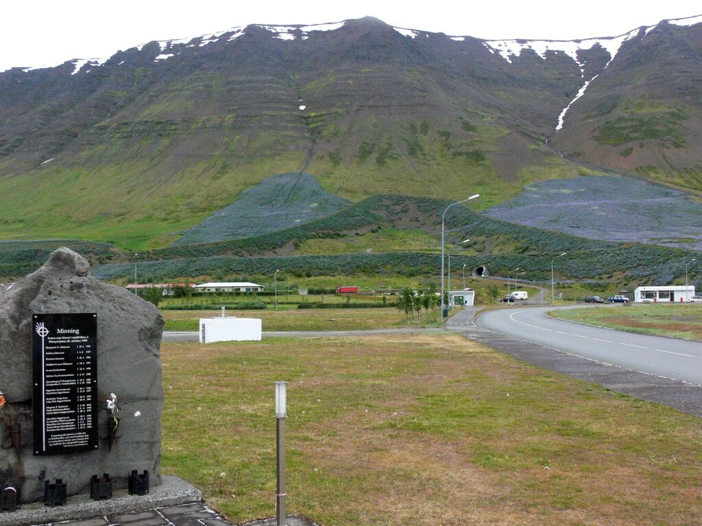 Memorial stone to the victims of the 1995 avalanche in Flateyri, with the lupine-covered A-shaped avalanche defense barriers visible on the mountainside behind the village.