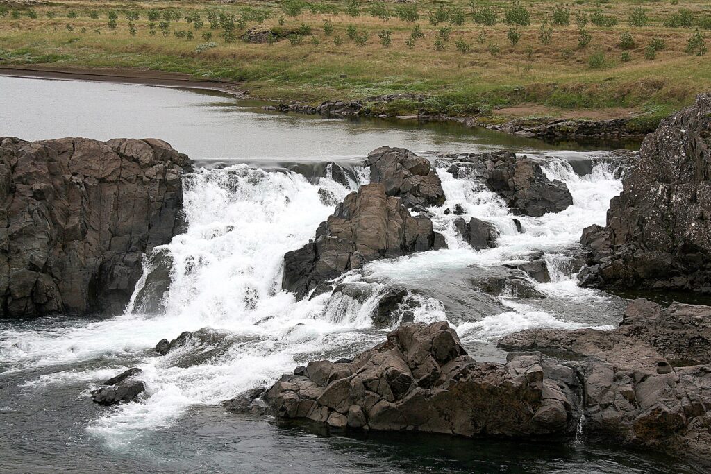 The Glanni waterfall spilling over dark lava ledges into the Norðurá river near Bifröst in West Iceland, with mossy green banks alongside.