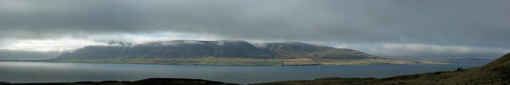 The calm waters of Hvalfjörður fjord in West Iceland, ringed by green and brown mountain slopes under a partly cloudy sky.