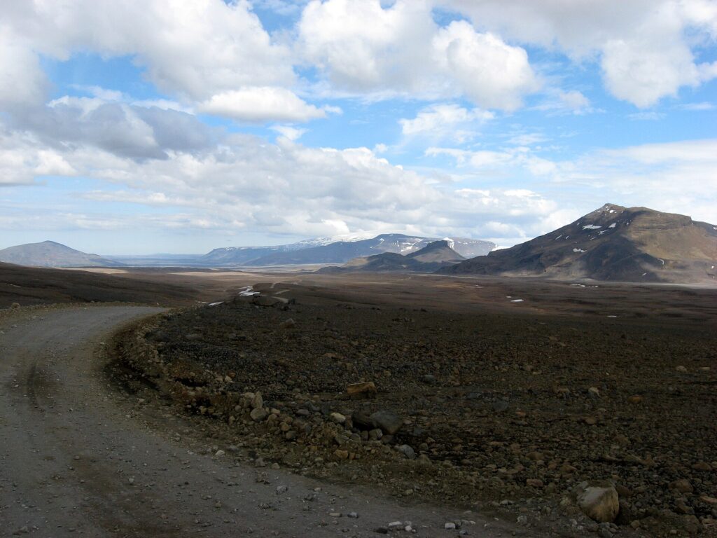 A gravel highland road heading north across the barren Kaldidalur valley in Iceland, with distant snow-streaked mountains and glacier under an overcast sky.