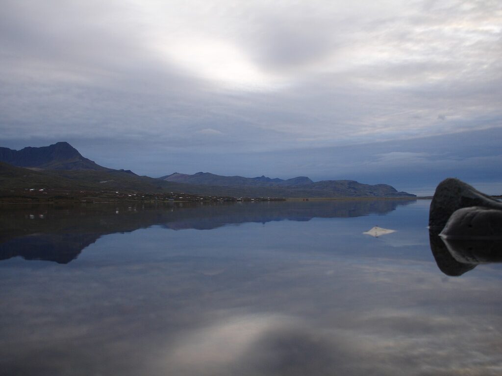 The pointed peak of Skessuhorn mountain reflected in the calm, dark water of Skorradalsvatn lake in West Iceland, with grassy shoreline in the foreground.